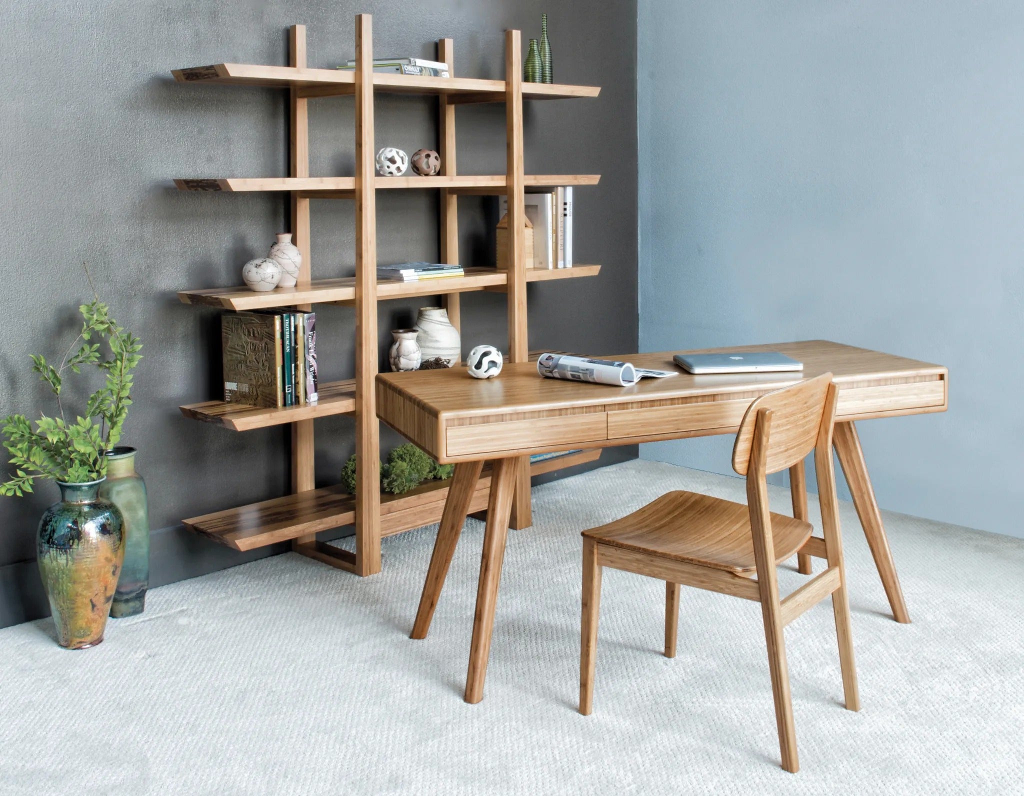 A renewable Currant Bamboo Writing Desk by Greenington in caramel color, placed in an office room, in front of a bookshelf and a chair.